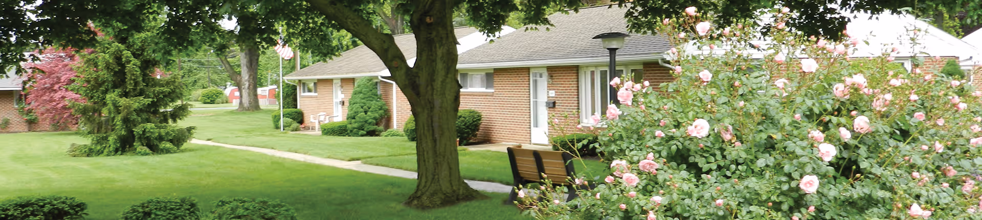 Low-rise brick senior living buildings with manicured lawns, trees, benches, and flowering bushes.
