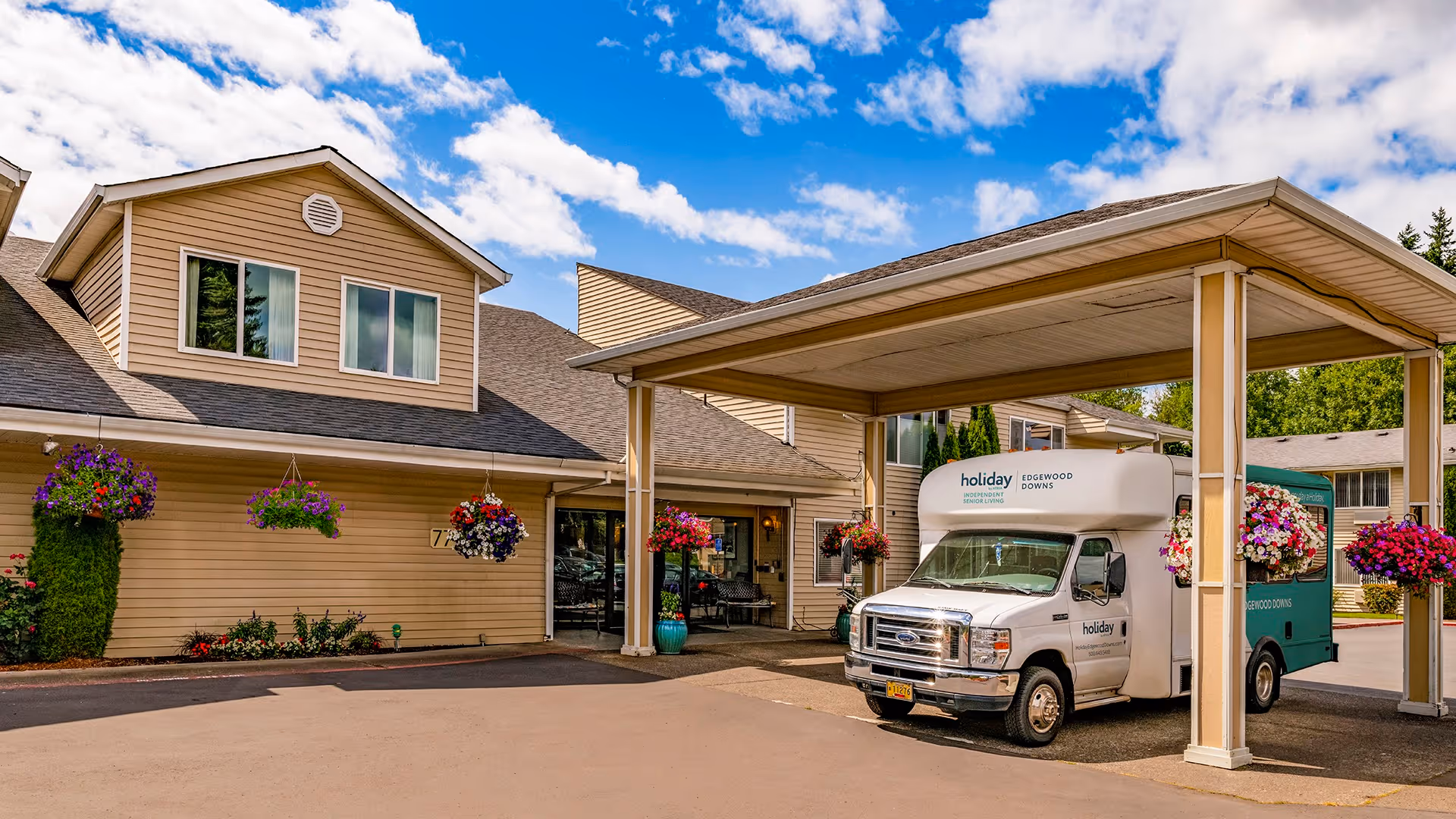 Exterior view of Holiday Edgewood Downs facility showing a beige building with hanging flower baskets and a covered entrance area where a white and green shuttle van is parked under the canopy.