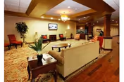 A spacious and warmly lit living room area with beige sofas, red chairs, a wooden coffee table, and a side table with a plant. The room features a patterned carpet, wooden flooring, decorative columns, and a flat-screen TV mounted on the wall.