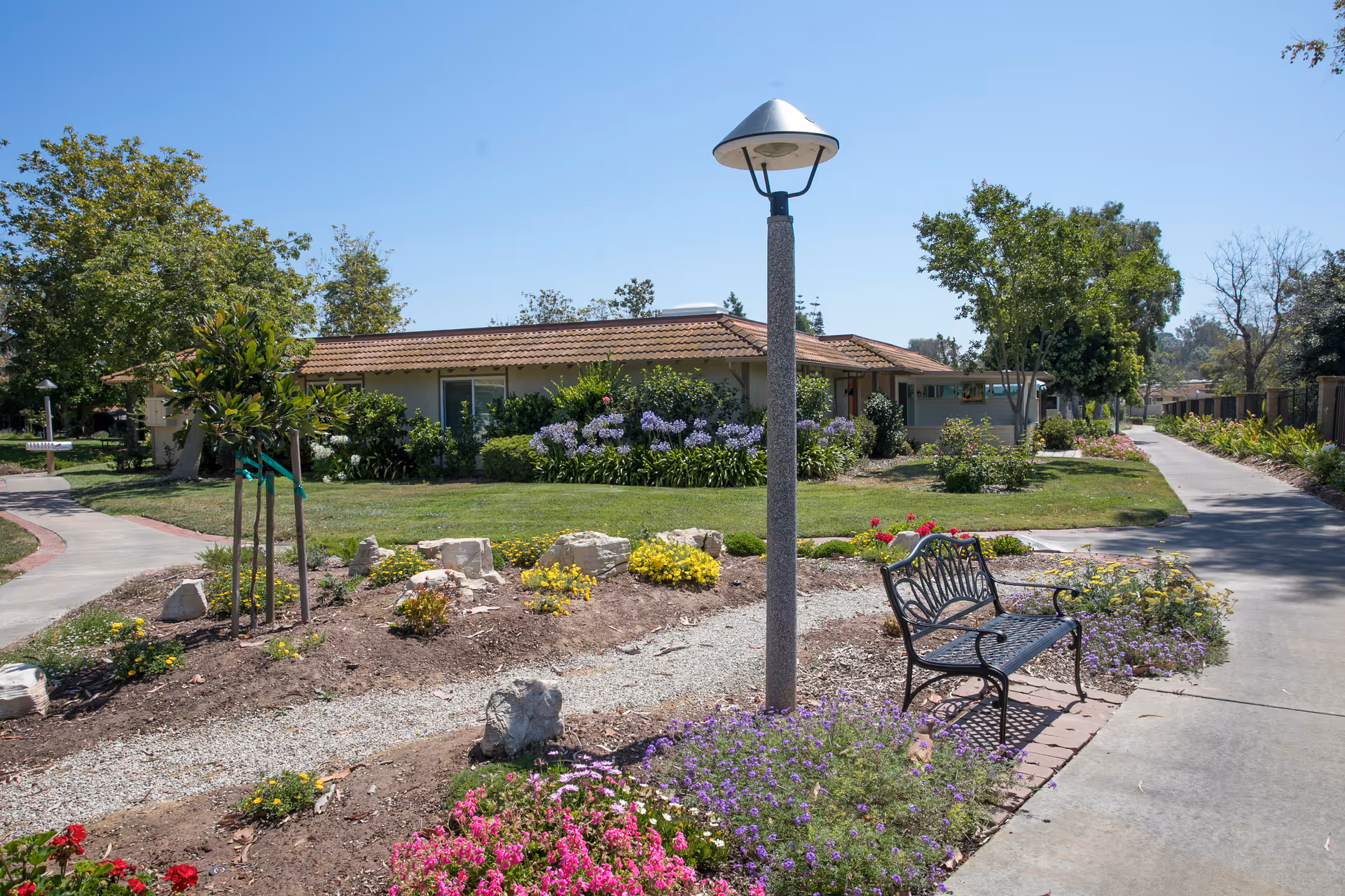 Outdoor garden area at The Willows senior living facility featuring a paved walkway, a black metal bench, a lamp post, various flowering plants, shrubs, and trees with a single-story building with a tiled roof in the background under a clear blue sky.