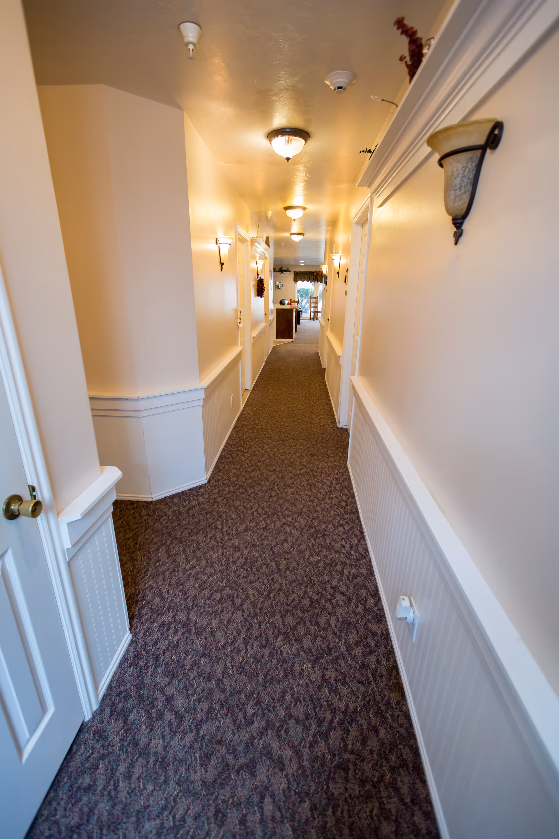 Long carpeted corridor in an assisted living facility lined with wainscoting, wall sconces, and doorways.
