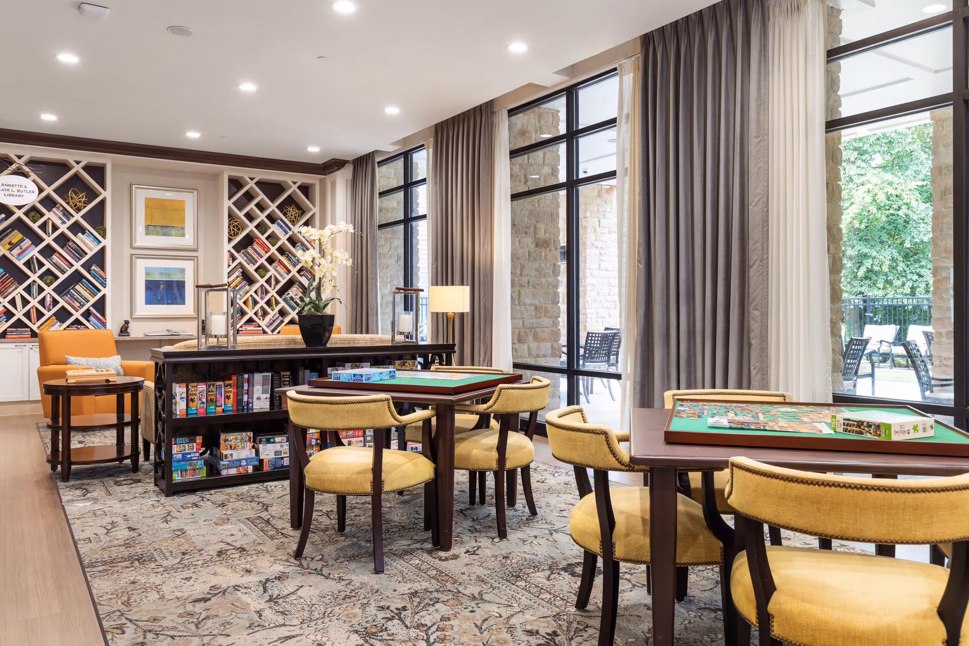 A bright and spacious common area in an assisted living facility featuring two tables with yellow cushioned chairs arranged around them. The tables have board games set up on top. Behind the tables is a bookshelf filled with various board games and books. The room has large windows with beige and gray curtains, allowing natural light to illuminate the space. There is also a comfortable orange armchair and framed artwork on the wall, creating a cozy and inviting atmosphere.