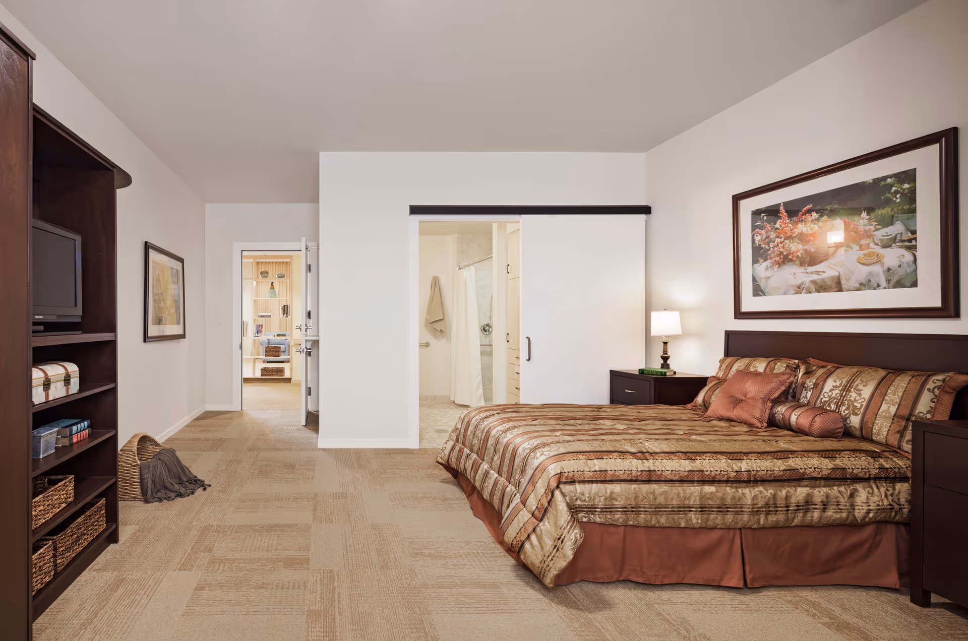 Spacious bedroom with a large made bed, nightstand and lamp, shelving unit, and open doorway to a bathroom.