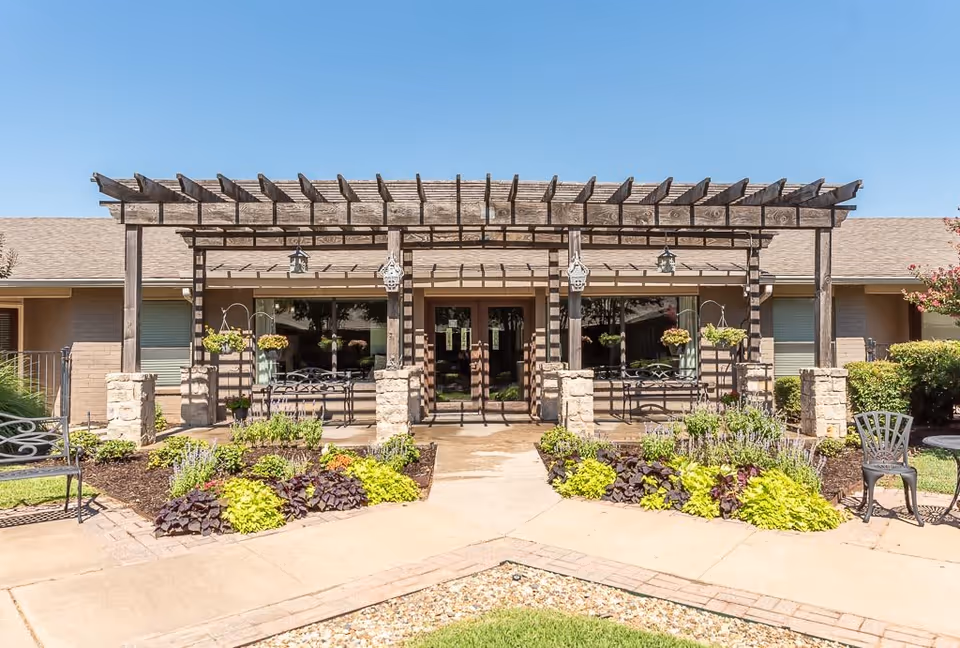 Outdoor view of a senior living facility entrance with a wooden pergola supported by stone pillars. There are hanging flower pots, benches, and well-maintained garden beds with various plants and shrubs. The building has large windows and a double glass door under the pergola, with a clear blue sky overhead.