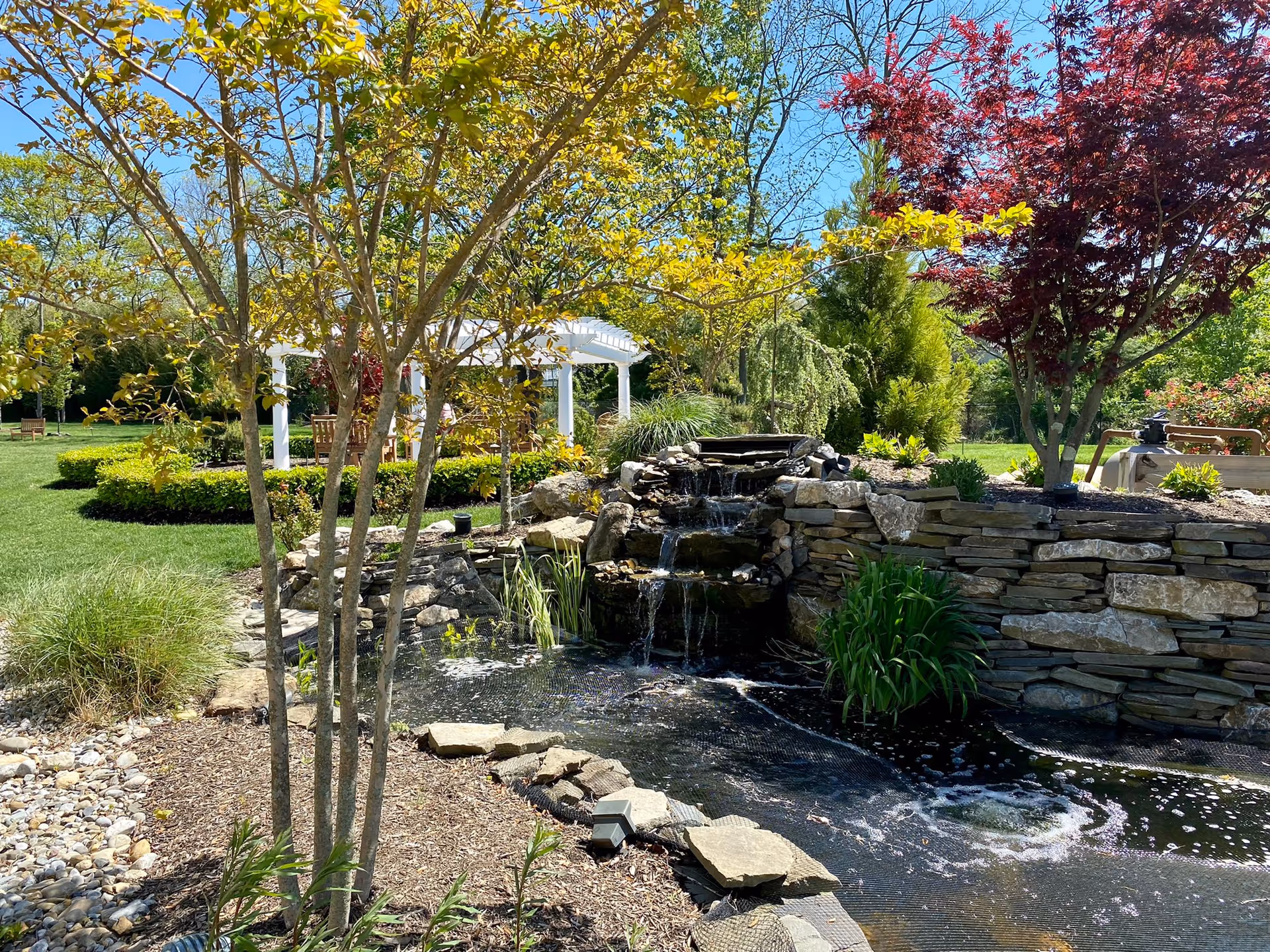 A landscaped outdoor garden area featuring a small cascading waterfall over stone steps into a pond, surrounded by various trees and shrubs. In the background, there is a white pergola with wooden benches underneath, set on a well-maintained green lawn under a clear blue sky.