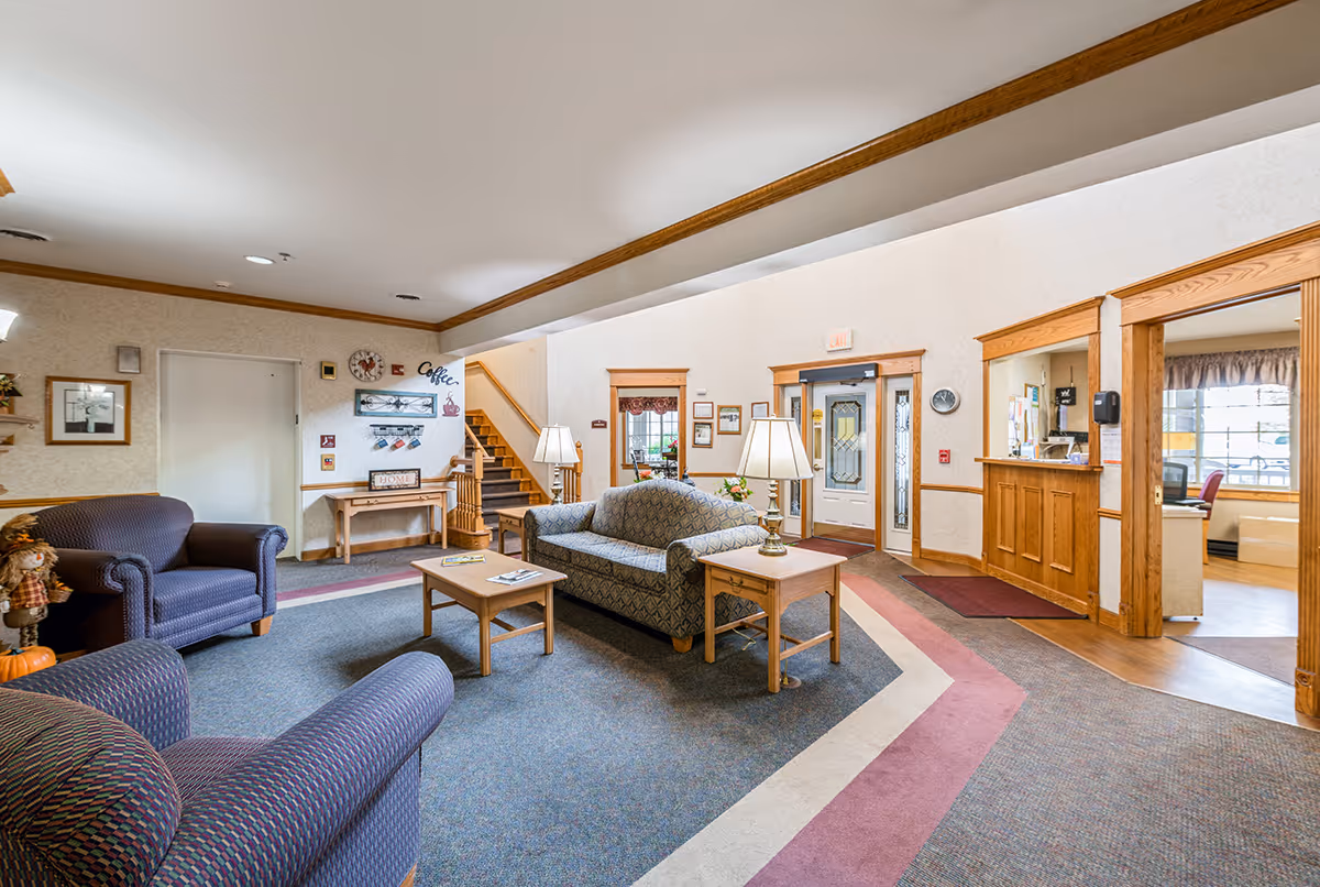 A cozy senior living facility common area with patterned armchairs and a sofa around wooden coffee and side tables with lamps. The room has carpeted flooring with a geometric pattern, wood trim, and a reception desk area to the right. There is a staircase in the background and a door with glass panels leading outside.