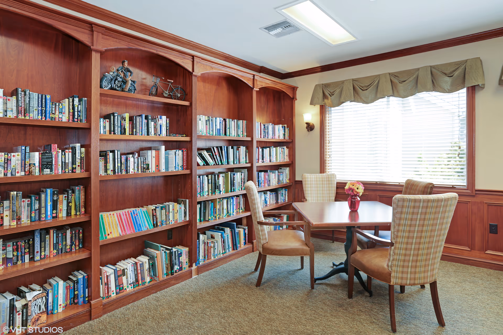 Cozy library-style common room with wooden bookshelves, a table with four chairs, and a window with blinds.