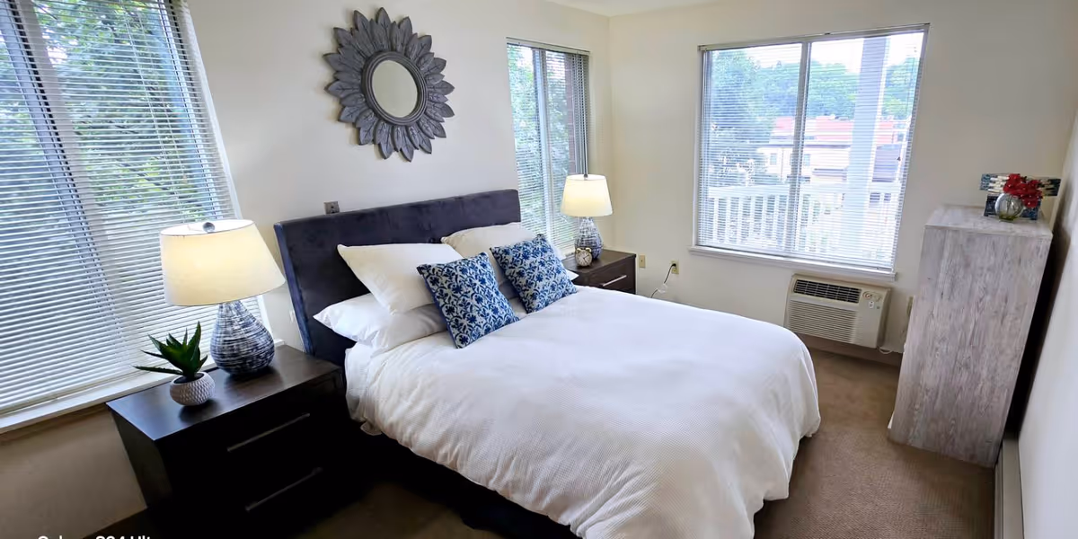 Bright furnished bedroom with a bed topped by decorative pillows, two nightstands with lamps, large windows, and a dresser.