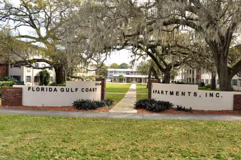 Exterior view of Florida Gulf Coast Apartments entrance with signs, walkway, and towering live oaks.
