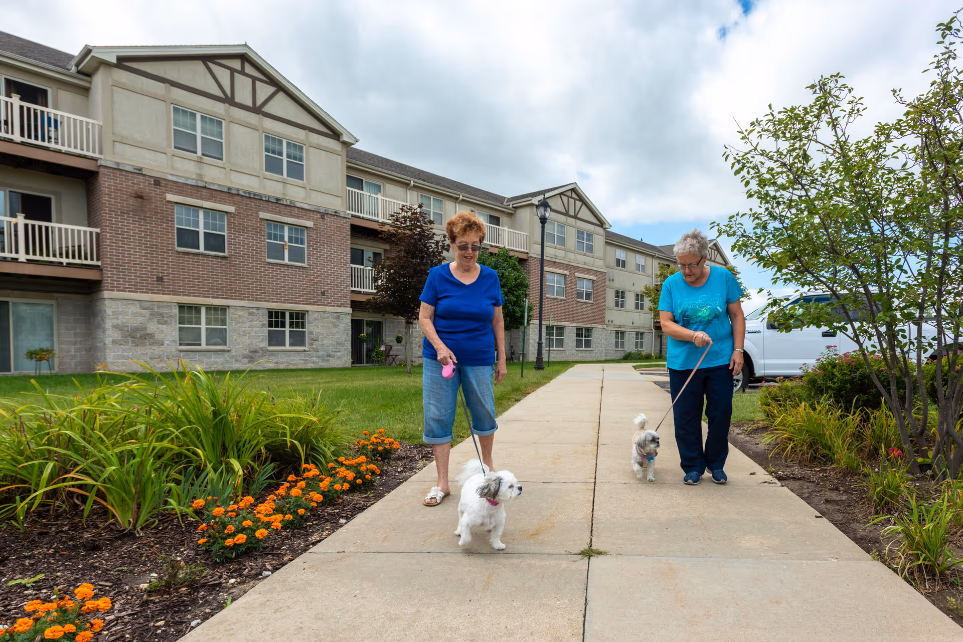 Two older women walking small white dogs on a sidewalk in front of a three-story senior living building with landscaped flower beds.