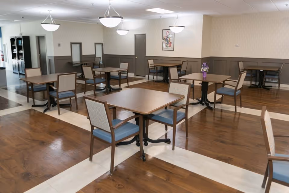 A clean and well-lit dining room with multiple square wooden tables and cushioned chairs arranged neatly on a wooden floor with light-colored tile accents. The walls are decorated with wainscoting and a framed artwork, and there are ceiling lights providing illumination.
