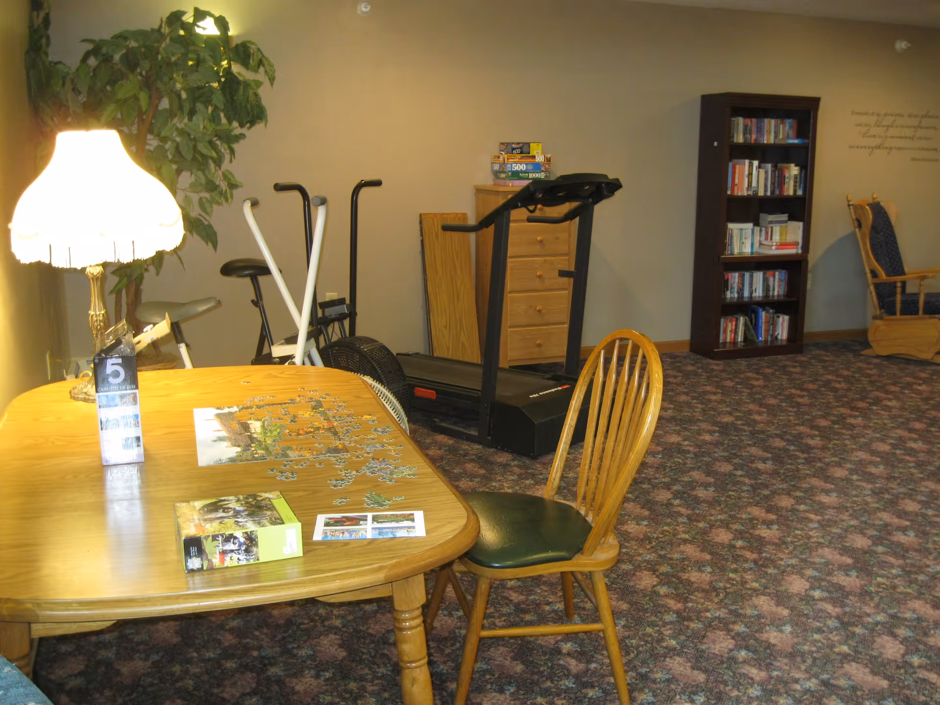 A cozy room with a wooden table and chair in the foreground, a partially completed jigsaw puzzle on the table, a lamp with a white lampshade, exercise equipment including a treadmill and stationary bike, a bookshelf filled with books, and a wooden rocking chair against the wall. The room has patterned carpet and beige walls.