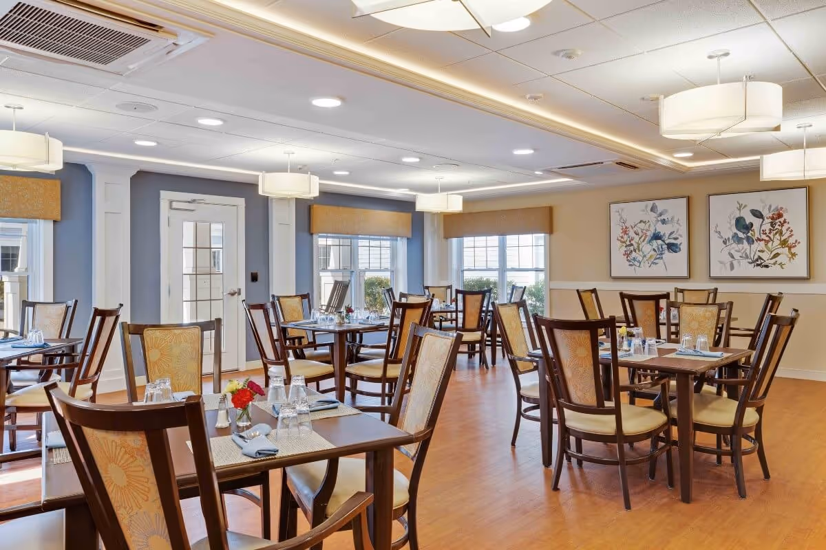 Bright dining room with multiple wooden tables and chairs set with place settings beneath pendant lights, windows, and floral wall art.