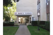 Entrance of a brick apartment building with a canopy sign reading 'St. Mark's Terrace', a concrete walkway, trimmed hedges and lamp posts.
