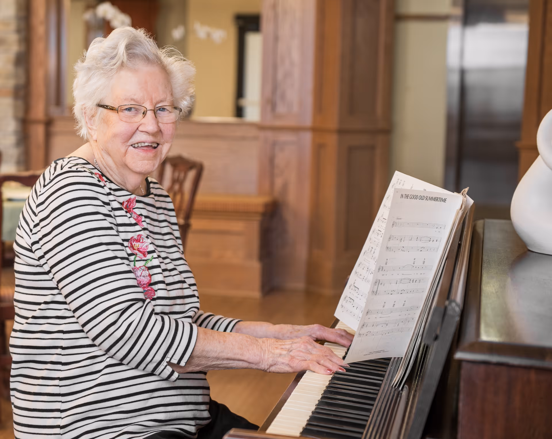 An elderly woman with white hair and glasses is smiling while playing a piano. She is wearing a striped shirt with floral embroidery. Sheet music titled 'In the Good Old Summertime' is placed on the piano. The background shows a warm, wood-paneled interior.