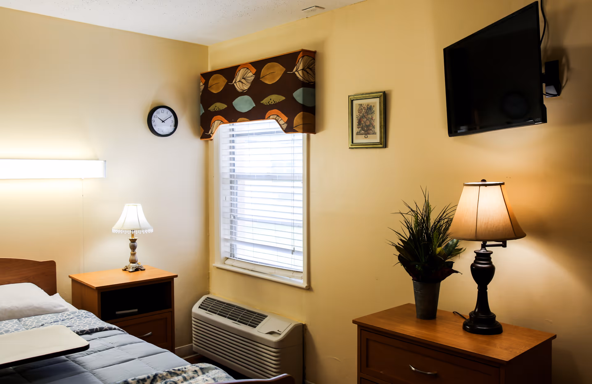 A cozy bedroom with a single bed, two wooden nightstands with lamps, a wall-mounted TV, and a window with blinds and an air conditioner below.