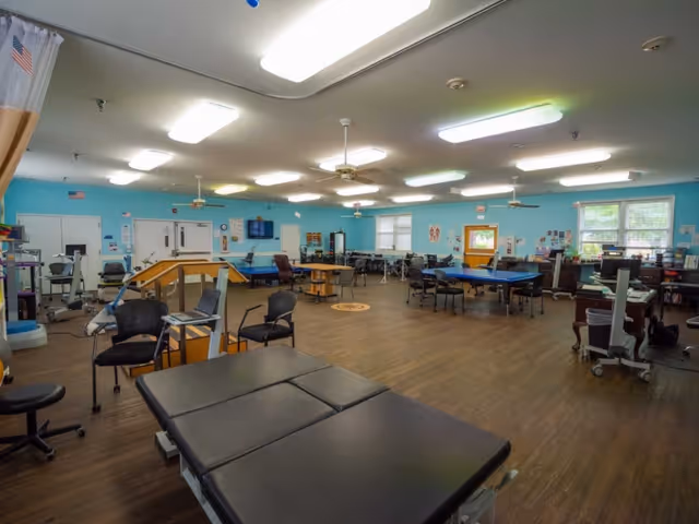 A spacious rehabilitation therapy room with light blue walls and wood flooring. The room contains various therapy equipment including treatment tables, chairs, parallel bars, exercise machines, and therapy tables. Ceiling fans and fluorescent lights are mounted on the ceiling, and windows allow natural light to enter the room.