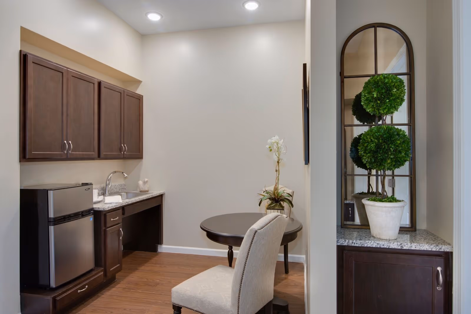 Small kitchenette with dark wood cabinets, a mini refrigerator and sink, a round table with a chair, and a mirrored niche holding a potted topiary.