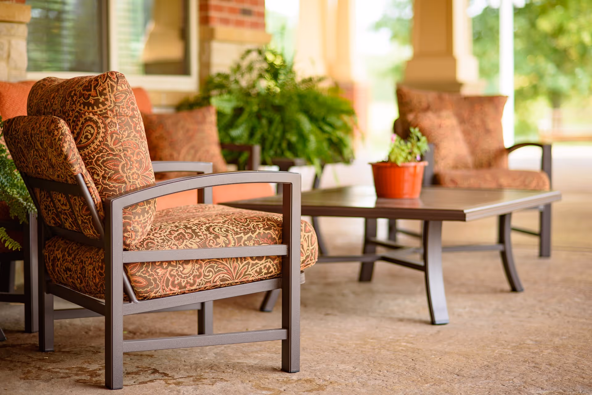 Outdoor seating area with cushioned armchairs featuring patterned upholstery and a rectangular coffee table with a small potted plant on top, set on a stone floor with greenery and brick pillars in the background.