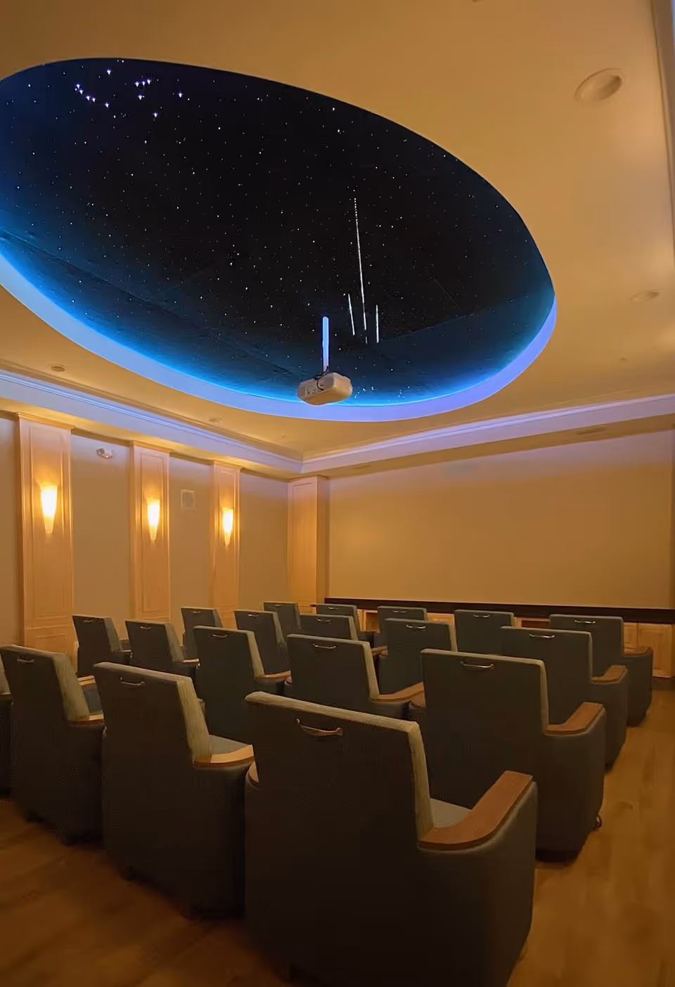 A small theater room with several rows of comfortable armchairs facing a blank wall, featuring a ceiling designed to look like a starry night sky with a projector mounted in the center.