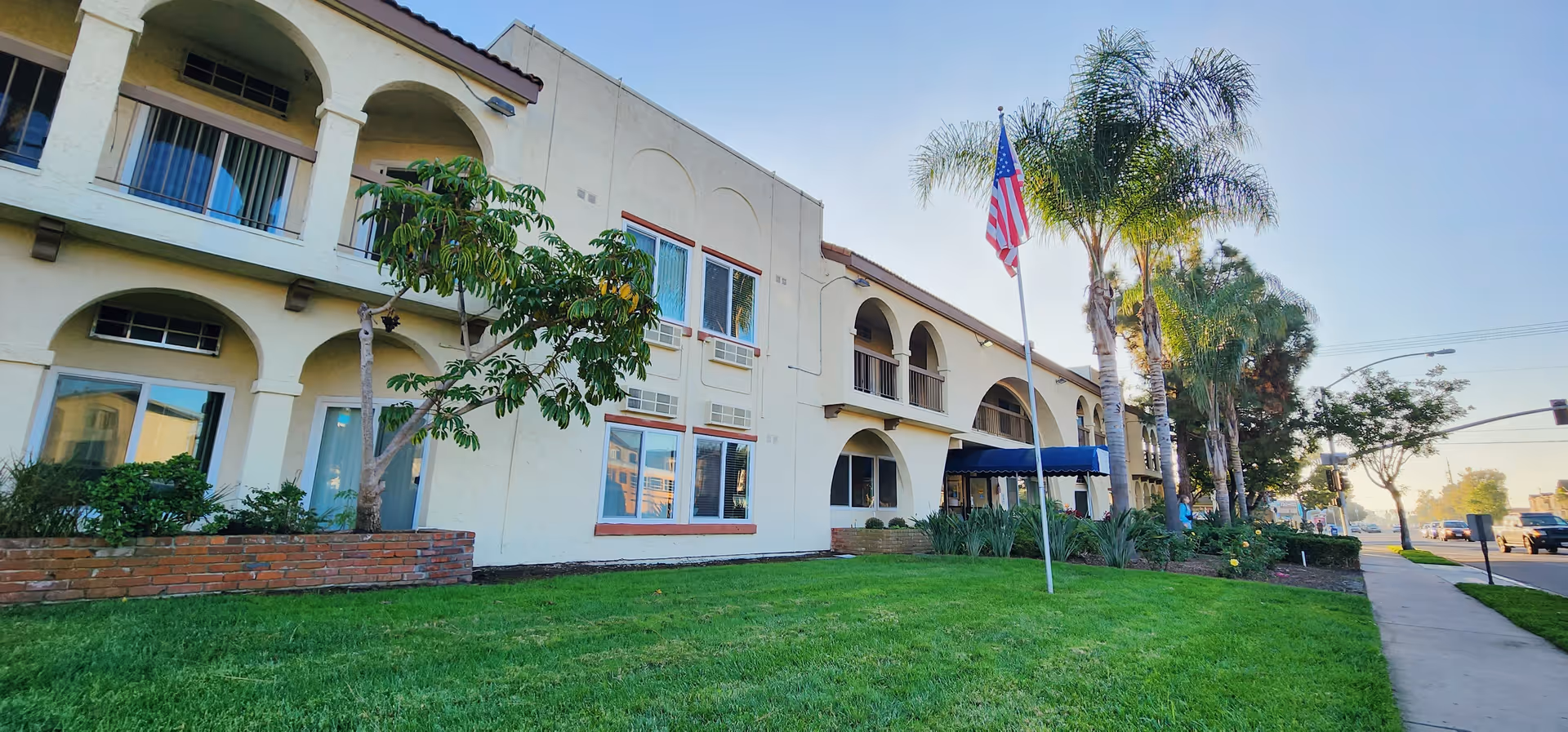 Front exterior of a two-story beige senior living building with arched balconies, palm trees, an American flag, and a grassy lawn.