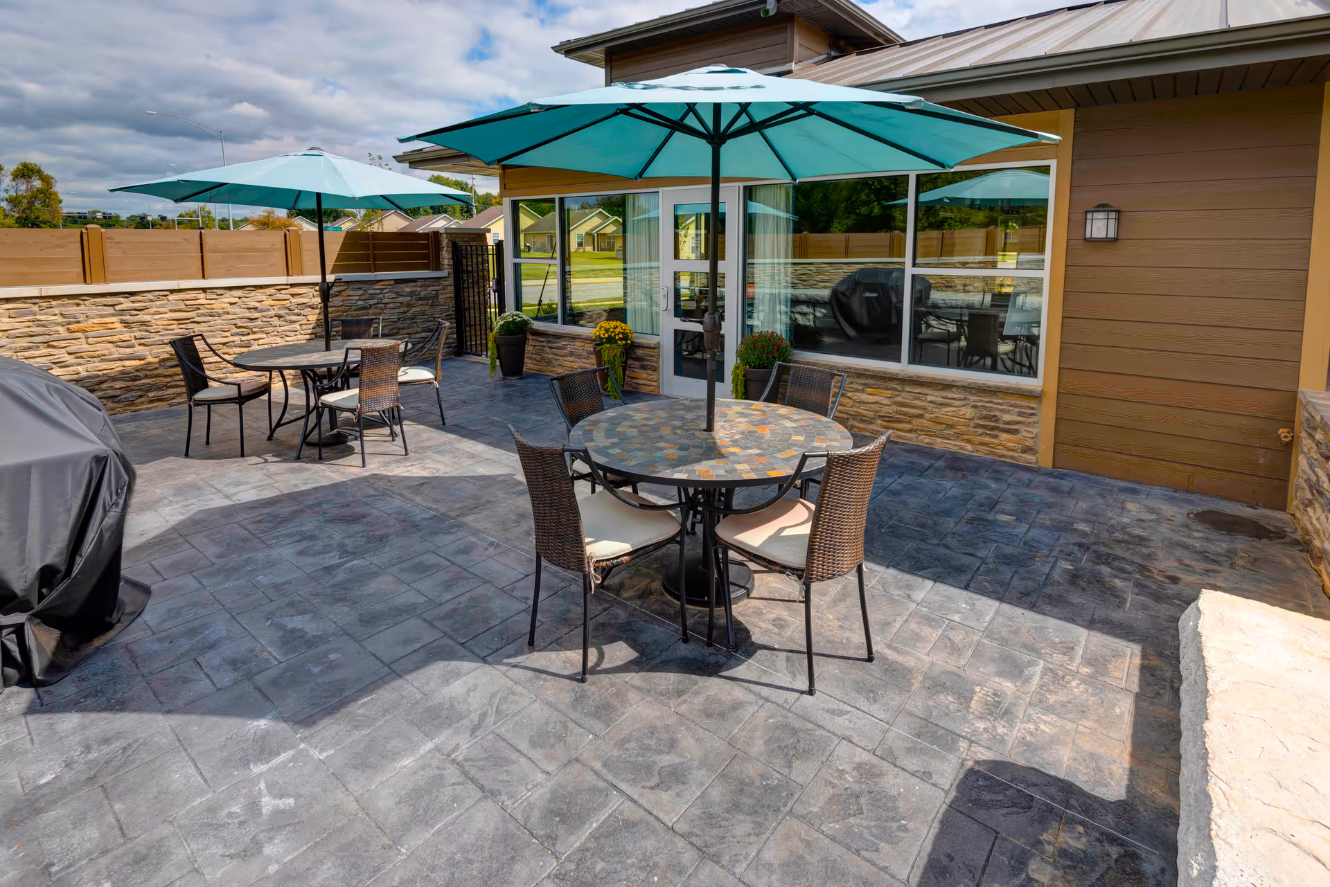 Outdoor patio area with two round tables, each with four chairs and large teal umbrellas. The patio is paved with stone tiles and surrounded by a stone and wood fence. A building with large windows and a door is visible in the background, along with some potted plants and a covered grill.