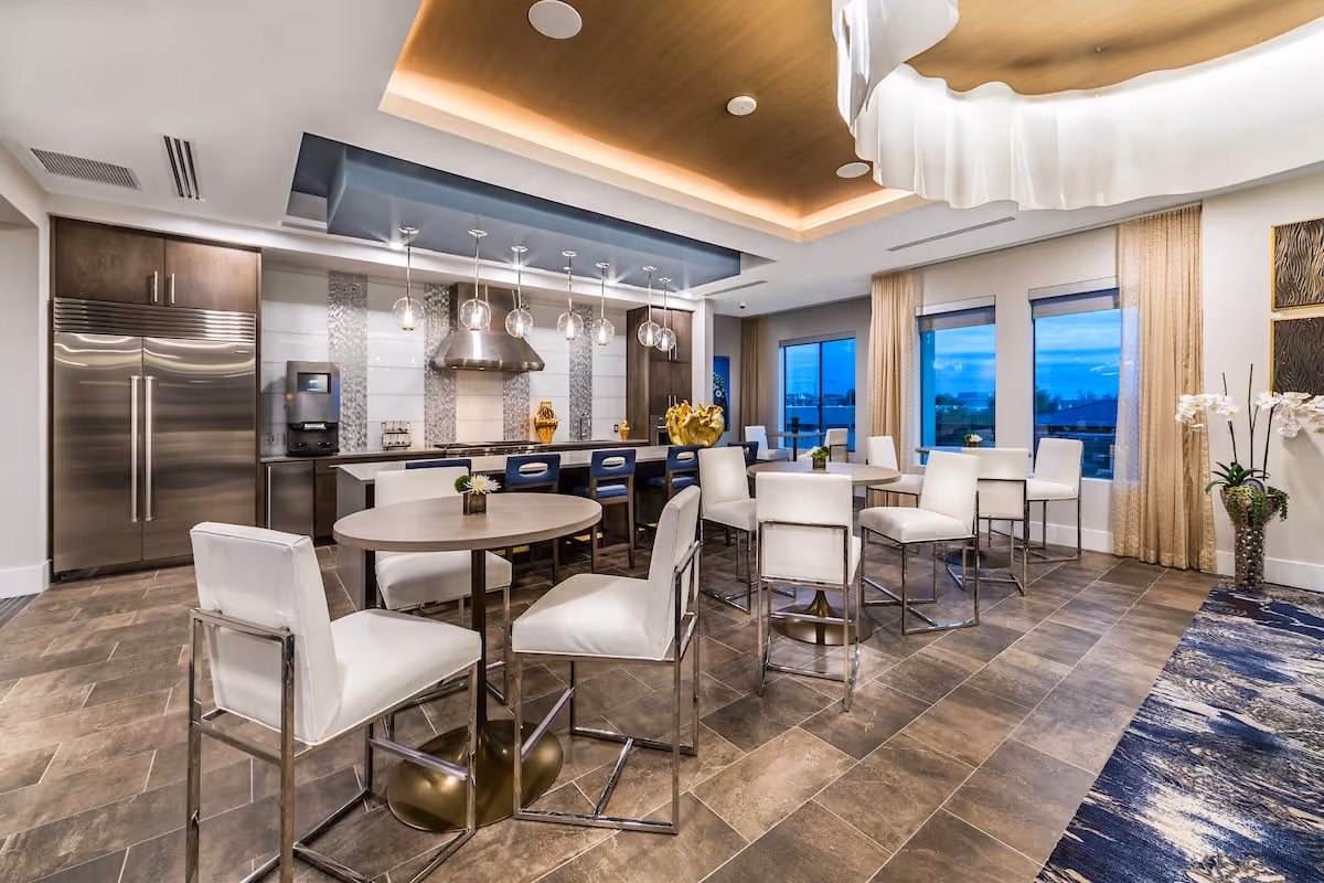 A modern dining area with multiple round tables and white cushioned chairs. The space features a large kitchen area with stainless steel appliances, including a double-door refrigerator and a range hood. Pendant lights hang above the kitchen island with bar stools. Large windows with beige curtains provide natural light and a view outside. The floor is tiled and there is a decorative rug on one side.