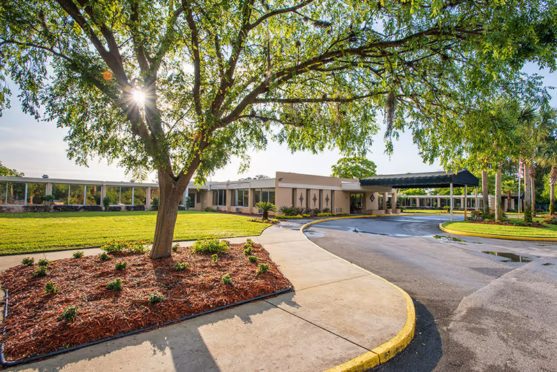 Front entrance of a single-story rehabilitation center with a circular drive, covered drop-off canopy and landscaped lawn under a large tree.