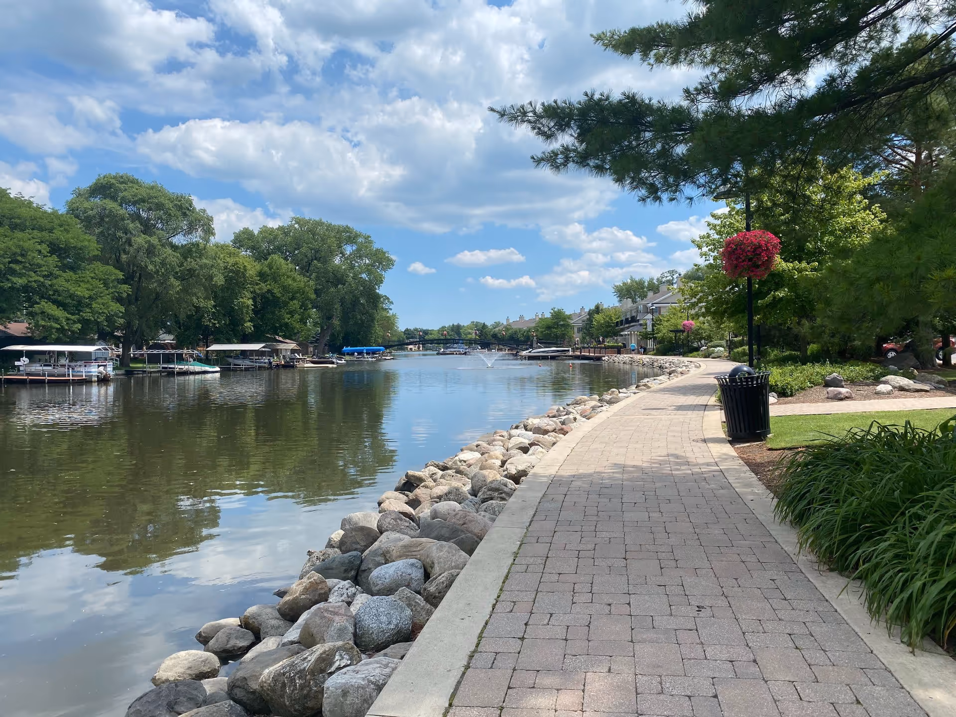 A paved walkway along a calm river with rocks lining the riverbank. Trees and hanging flower baskets are visible along the path, with houses and boats docked on the opposite side of the river under a partly cloudy blue sky.