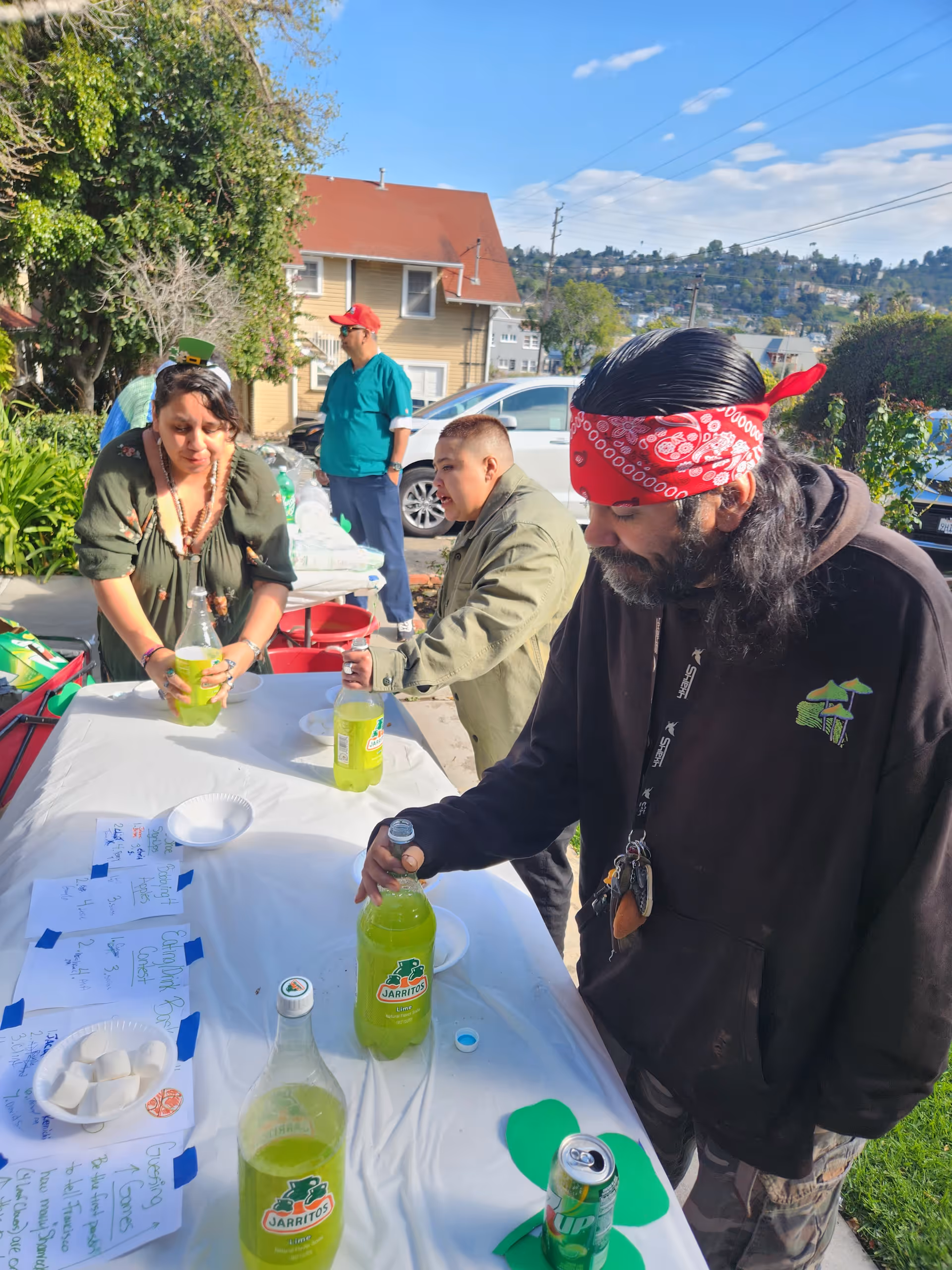 Three people standing around a table outdoors on a sunny day, each holding or opening bottles of Jarritos lime soda. The table is covered with a white cloth and has several bottles of soda, paper plates, and handwritten notes. In the background, there are houses, cars, trees, and a hill under a partly cloudy sky.