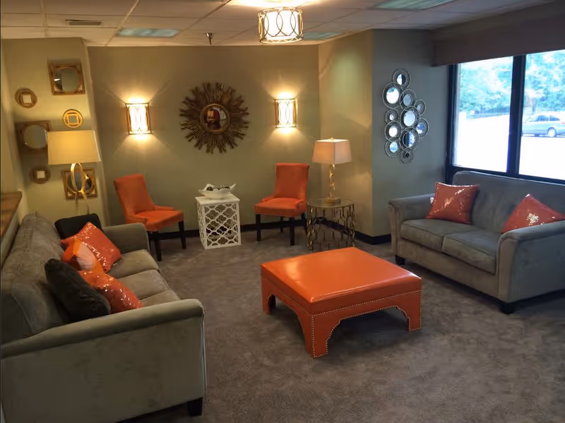 A cozy living room with two gray sofas adorned with orange and black pillows, two orange chairs, a large orange ottoman in the center, two table lamps, decorative mirrors on the walls, and a large window letting in natural light.