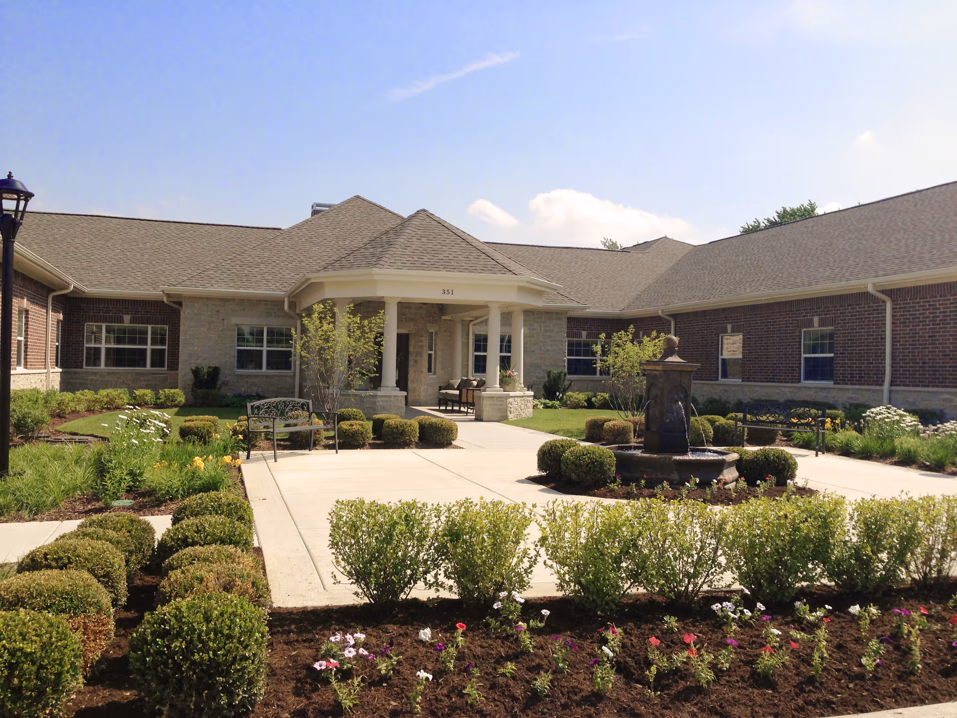 Entrance courtyard of a single-story senior living facility with a central fountain, benches, and manicured flowerbeds.