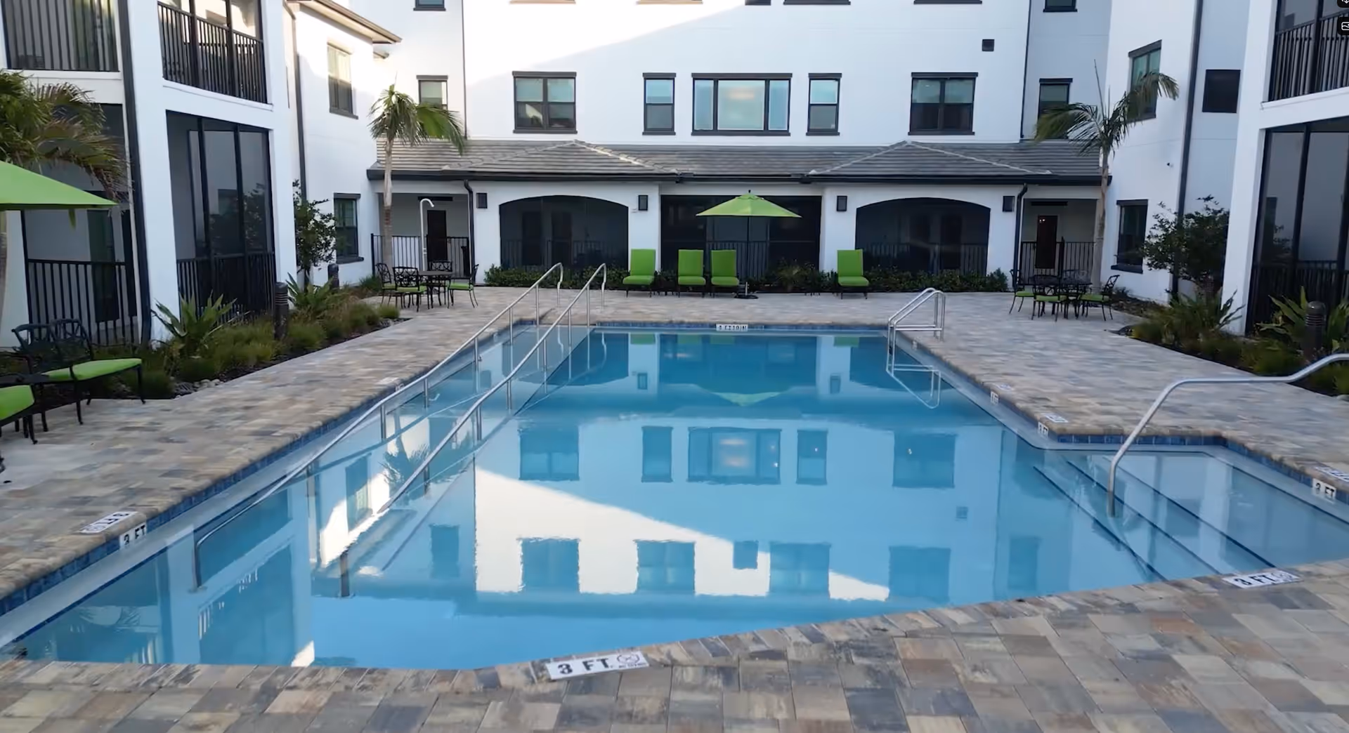 Outdoor swimming pool in the courtyard of a senior living facility with lounge chairs, tables, umbrellas, and surrounding white multi-story building.