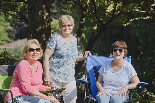Three elderly women wearing sunglasses enjoying a sunny day outdoors, sitting and standing near folding chairs with trees and greenery in the background.