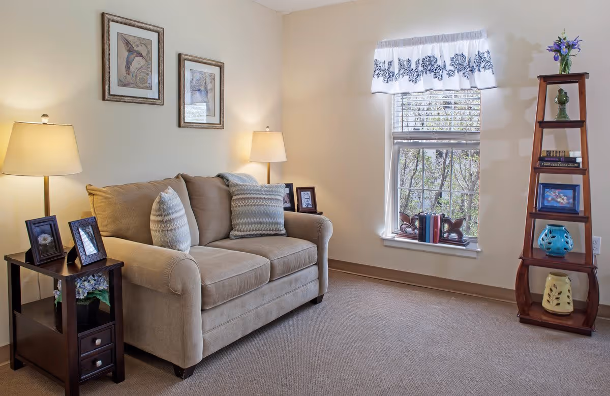 Cozy living room with a beige loveseat, side table with lamps and framed photos, a window with a valance, and a wooden ladder-style shelving unit with decor.