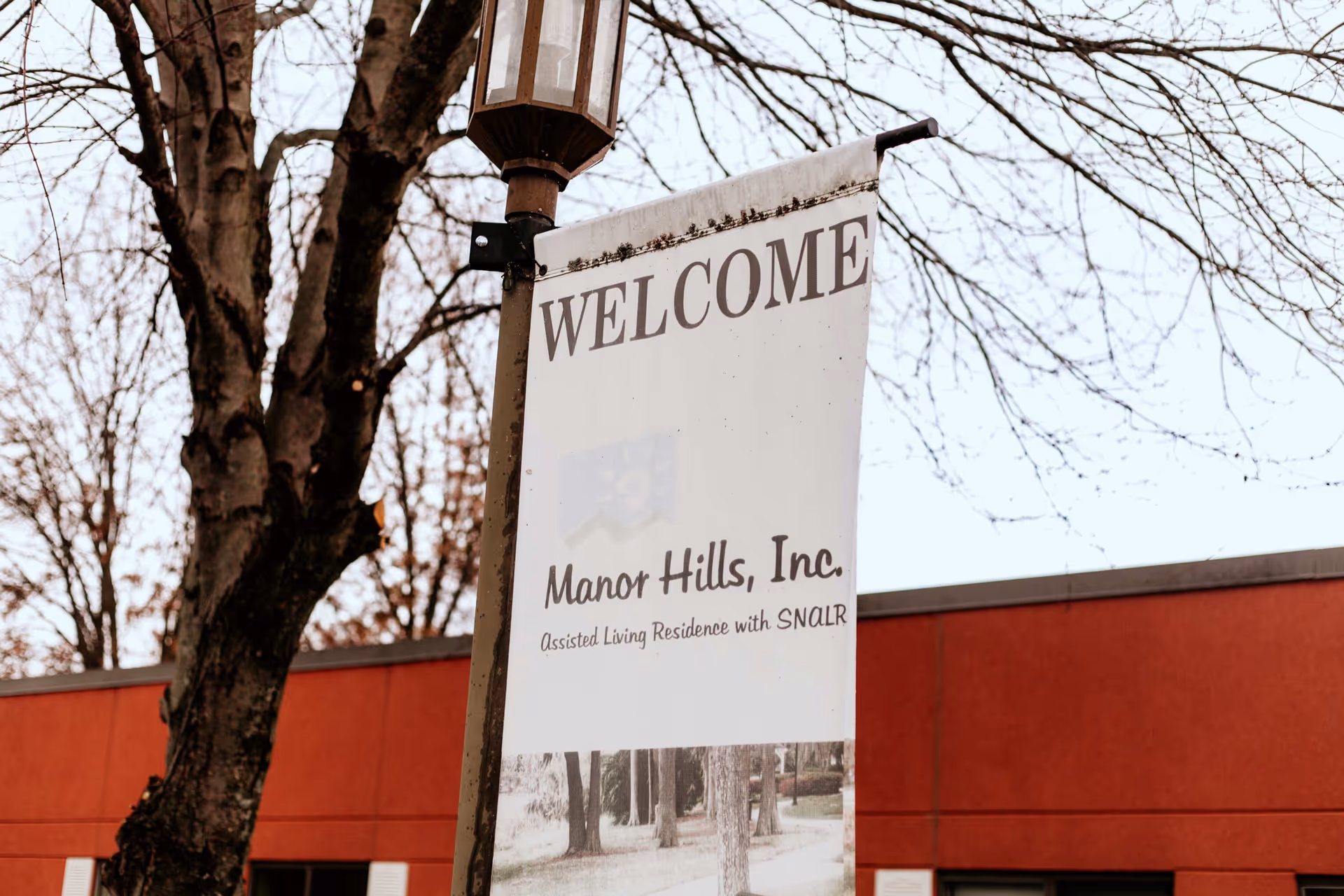 A white vertical banner hanging from a street lamp post with the text 'WELCOME Manor Hills, Inc. Assisted Living Residence with SNALR'. Behind the banner, there is a tree with bare branches and a red building with windows.