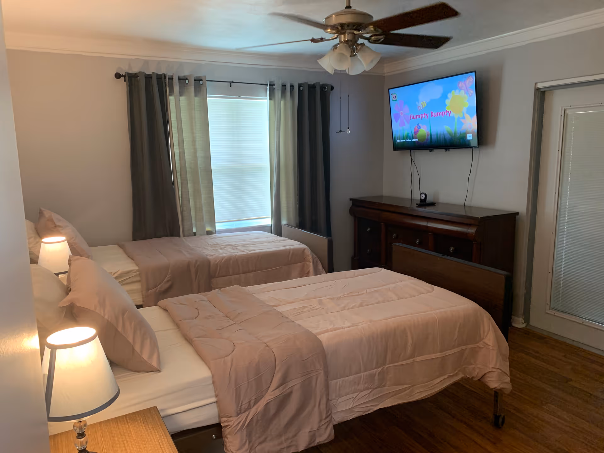 Two neatly made twin beds in a bedroom with bedside lamps, curtains, a ceiling fan, a dresser and a wall-mounted TV.