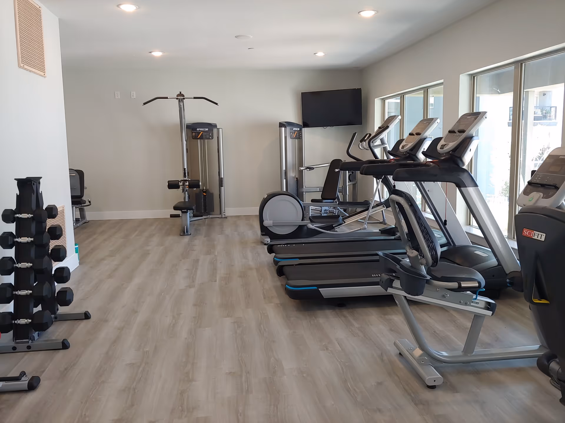 A fitness room with exercise equipment including treadmills, a recumbent bike, weight machines, and a rack of dumbbells. The room has light wood flooring, white walls, recessed ceiling lights, and large windows letting in natural light. A flat-screen TV is mounted on the wall.