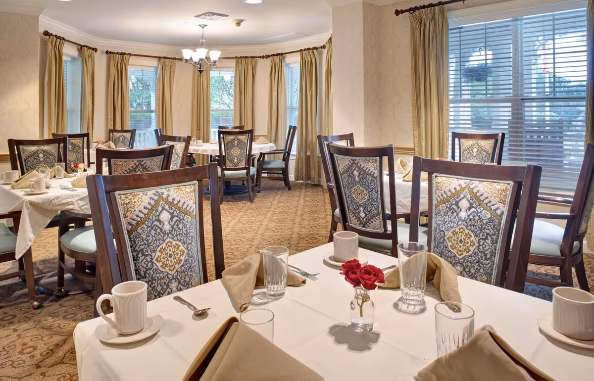 Dining room with tables set for a meal, upholstered chairs, place settings and a small vase of red roses by curtained windows.