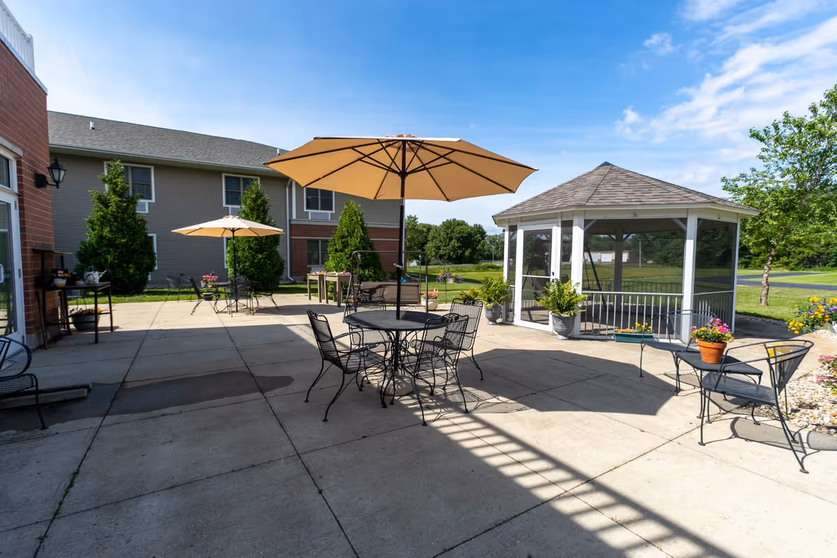 Sunlit outdoor patio with metal tables and chairs, umbrellas, potted plants, and a screened gazebo in front of the facility building.