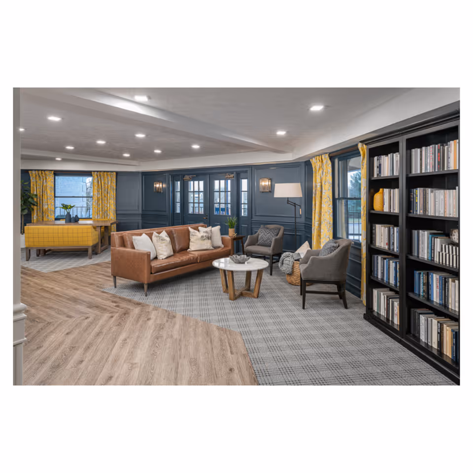 Cozy communal lounge with a brown leather sofa, gray armchairs, a round coffee table, bookshelf, and yellow curtains.