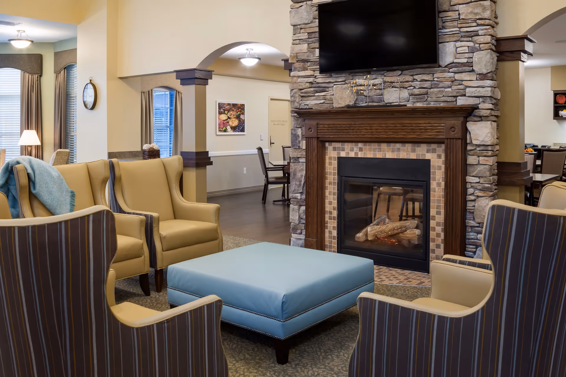 A cozy living room area with a stone fireplace and a mounted flat-screen TV above it. The room features four armchairs arranged around a large blue ottoman. The walls are painted in warm tones, and there are windows with curtains letting in natural light. In the background, there is a dining area with tables and chairs.
