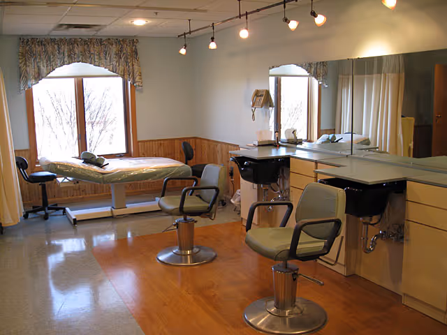A well-lit room with two salon-style chairs in front of a long mirror and two sinks, a treatment table near a window with floral curtains, and a telephone mounted on the wall. The room has wood paneling on the lower half of the walls and a wooden floor area in the center.