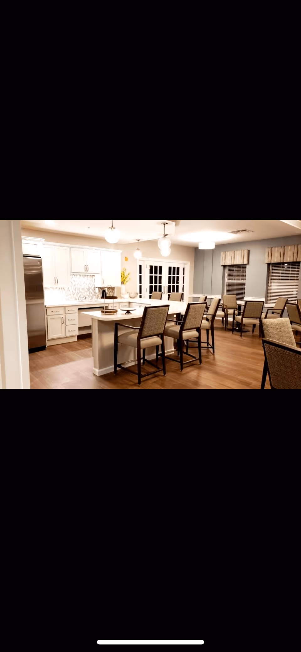 A spacious and well-lit kitchen and dining area with white cabinetry, stainless steel refrigerator, a large island with seating, and multiple tables and chairs arranged for dining. The room has wooden flooring, pendant lights, and windows with blinds and valances.