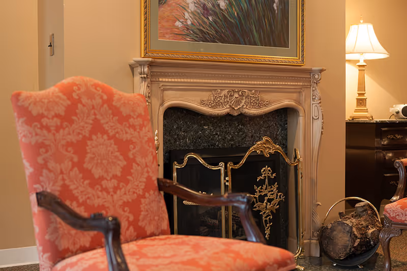 A cozy living room corner featuring an ornate fireplace with a decorative screen, a coral patterned upholstered armchair with wooden arms, a side table with a lamp, and a basket holding firewood. A framed painting hangs above the fireplace.