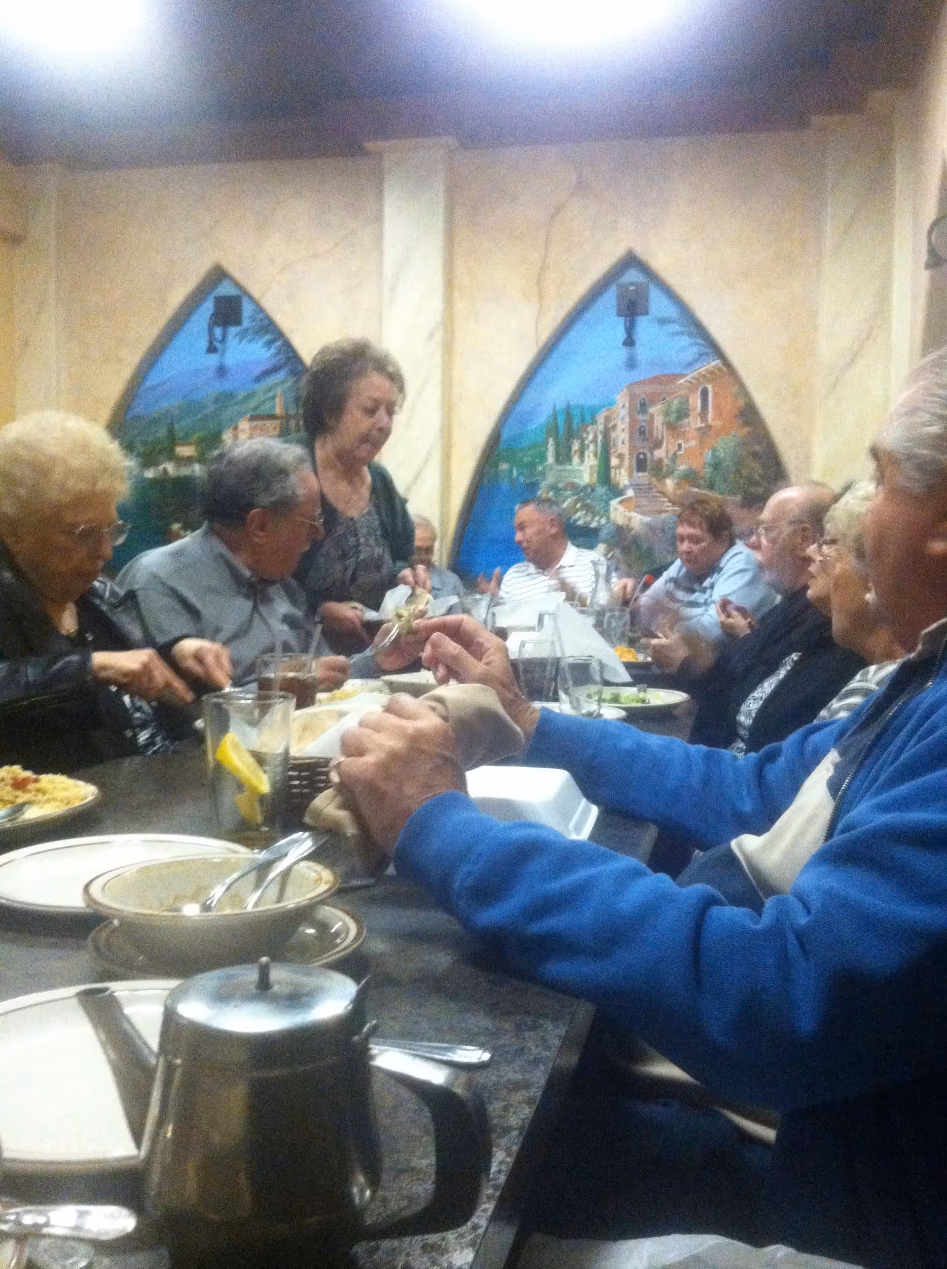 A group of elderly people sitting around a long dining table enjoying a meal together. The room has decorative wall art depicting scenic landscapes with buildings and water. There are plates, glasses, and a metal teapot on the table.