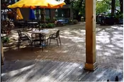 Outdoor patio area with a round table and four metal chairs under a large yellow umbrella. The patio is paved with bricks and surrounded by trees and greenery, with a wooden post visible in the foreground.
