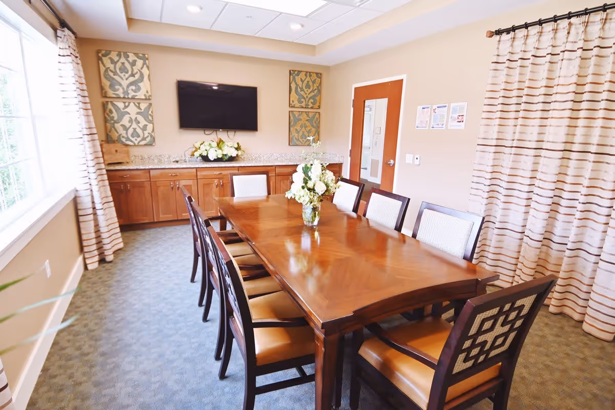 A well-lit dining room with a long wooden table surrounded by eight chairs. A vase with white flowers is placed in the center of the table. The room has large windows with striped curtains on one side and a wooden cabinet with a granite countertop along the back wall. Above the cabinet is a flat-screen TV mounted on the wall, flanked by two decorative wall hangings. A wooden door with a glass panel is visible on the right side of the room.
