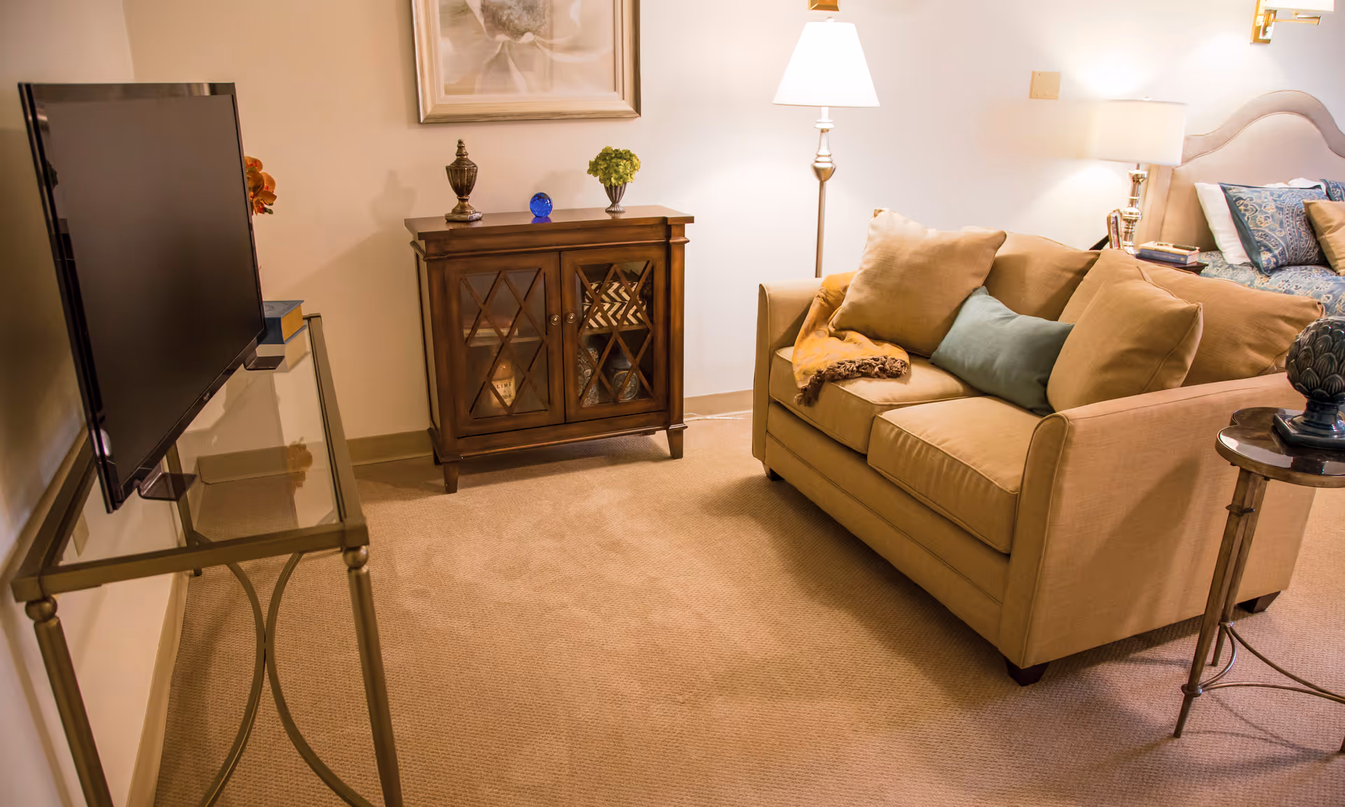 A cozy living room area with a beige loveseat adorned with multiple cushions and a throw blanket. To the left, a flat-screen TV is mounted on a glass and metal stand. Behind the loveseat, there is a wooden cabinet with glass doors displaying decorative items, and a floor lamp providing warm lighting. A side table with a decorative object is next to the loveseat, and a bed with pillows is partially visible in the background.