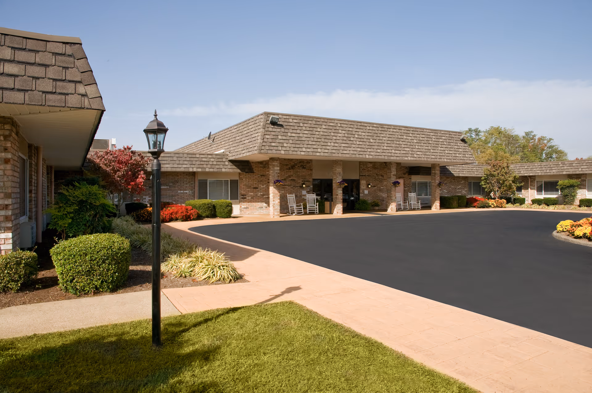 Exterior view of a single-story brick building with a shingled roof, a paved driveway, and landscaped bushes and flowers. There are rocking chairs near the entrance under a covered porch area.